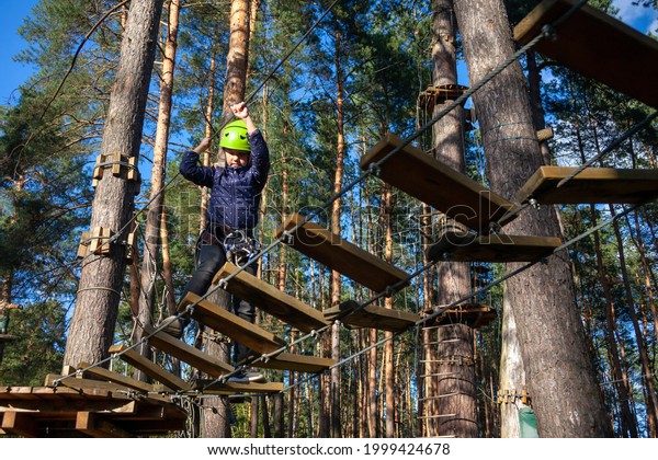 a girl in a green helmet with a safety net,\
smiling, runs along a rope bridge made of wooden steps a distance\
in a forest extreme rope park