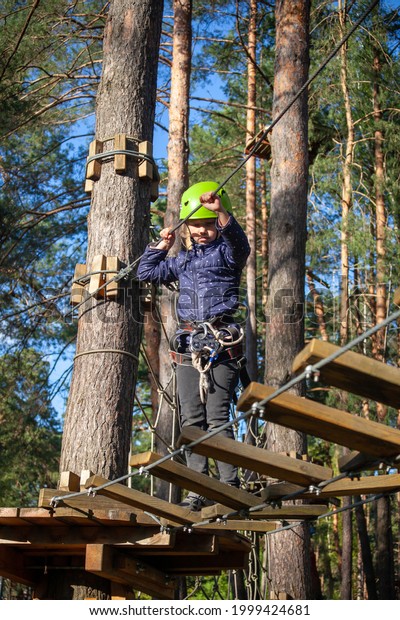 a girl in a green helmet with a safety net, smiling,\
runs along a rope bridge made of wooden bred a distance in a forest\
extreme rope park