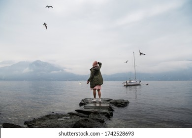 A girl in a green jacket and a pink hat stands with her back on the lake and enjoys the view. Spectacular view of Lake Como. Cloudy and rainy weather, seagulls fly in the sky