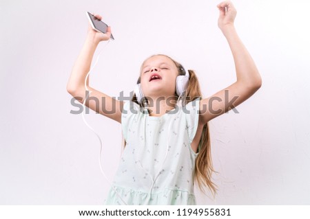 A girl in a green t-shirt listens to music from a smartphone in large white headphones and sings along on a white background.