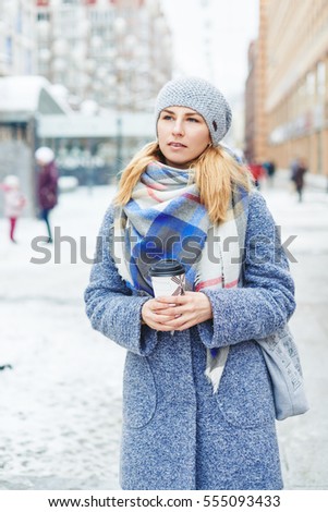 Girl in grey coat, hat and blanket scarf with cup of coffee on winter street, winter street style lifestyle
