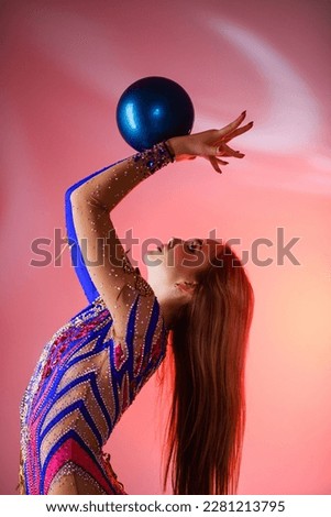 Girl gymnast wears a gymnastic leotard practicing with a ball on a pink background.