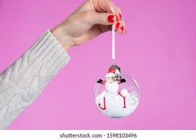 Girl hand holding ornament ball with snow isolated on pink background in studio. Female hands with red nails polish on fingers. Christmas or Valentines day holiday and minimalism concept