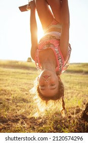 The Girl Is Hanging Upside Down On A Tree In Summer In Orange Sunlight And A Light Dress. Summer Time, Heat, Childhood. Funny Portrait With Disheveled Hair