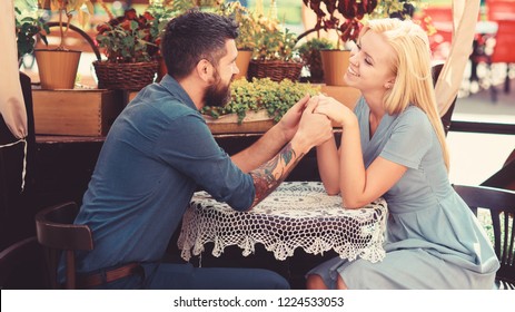 Girl happy and fall in love with bearded man, looks at him with adoration. Woman smitten with man, couple holding hands. Couple in love sit in cafe outdoor, urban background. Date and love concept.
