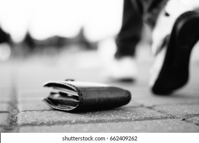 Girl has lost her wallet with money on city street. Close-up shot of brown leather wallet laying at sidewalk, and feet of woman going away on the background. Concept of money loss. Monochrome