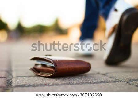 Girl has lost her wallet with money on city street at sunny summer day. Close-up shot of brown leather wallet laying at sidewalk, and feet of woman going away on the background. Concept of money loss
