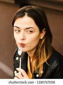 Girl Has Tasty Food On Brown Terrace Background. Woman With Tricky Face Stays At Cafe. Nutrition And Eating Out Concept. Lady Holds Dessert Fork In Mouth Looking Aside