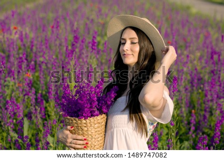 Girl with hat in a field of flowers, hold basket with flowers.Beautiful young woman with healthy hair over purple field landscape background. Attractive brunette girl with curly hair