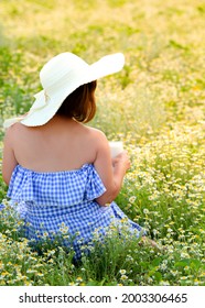 a girl in a hat is sitting among a field of daisies and drinking milk from a high transparent glass