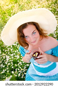 a girl in a hat is sitting on a chamomile field, drinking tea, in which chamomile inflorescences are floating, a beautiful girl is looking at the camera
