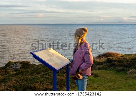Girl in hat standing next to information board looking out to sea 