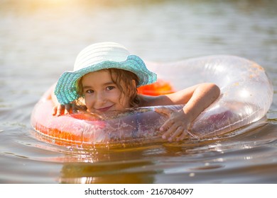 Girl in a hat stands on the river bank with a transparent inflatable circle in the shape of a heart with orange feathers inside. Beach holidays, swimming, tanning, sunscreens. - Shutterstock ID 2167084097