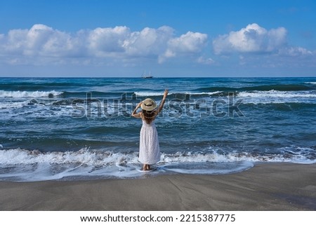 Girl with hat and white dress is standing near waves of Mediterranean Sea, waving hand goodbye at sailing vessel at the distance. Photo taken at Patara Beach, Antalya, Turkey   