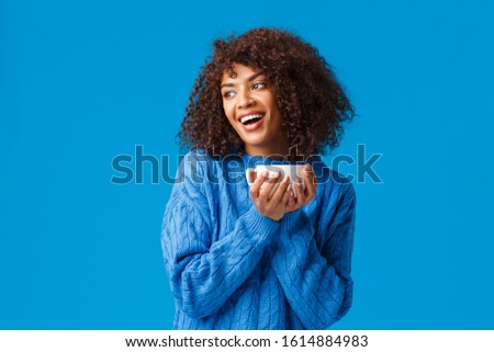 Girl having chat with girlfriend while drinking coffee. Cheerful and cute african-american lovely woman with afro haircut, in sweater, tilt head gazing left and holding hot cup delicious tea