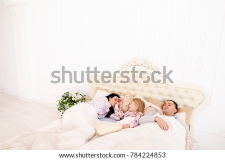 Girl having fun, teases parents nose, she wakes them and wants young couple woke up.Mom with black long hair, Dad European appearance, a daughter with long white hair smiling. Concept of happy family