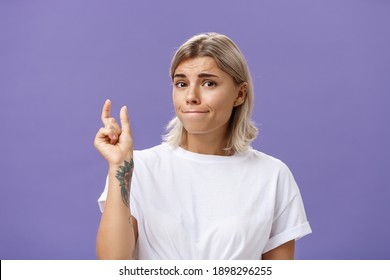 Girl Having Tiny Problem. Concerned Attractive Blonde Girl With Tattoo On Arm Pursing Lips In Troubled Look Shaping Small Or Little Object, Dissatisfied With Regret In Eyes Over Purple Wall