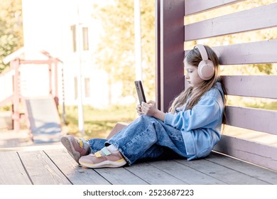 A girl with headphones listens to music on a smartphone outside. The young girl studies an online lesson, enjoys an audiobook, takes selfies and chats on social networks. - Powered by Shutterstock