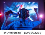 Girl in headphones plays a video game on the big TV screen. Gamer with a joystick. Online gaming with friends, win, prize. Fun entertainment. Teens play adventure games. Back view. Neon lighting