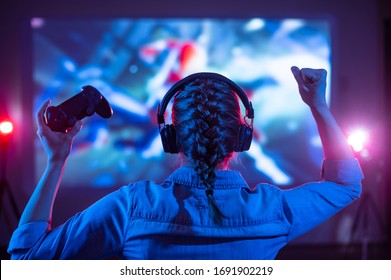 Girl in headphones plays a video game on the big TV screen. Gamer with a joystick. Online gaming with friends, win, prize. Fun entertainment. Teens play adventure games. Back view. Neon lighting - Shutterstock ID 1691902219