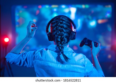 Girl in headphones plays a video game on the big TV screen. Gamer with a joystick. Online gaming with friends, win, prize. Fun entertainment. Teens play adventure games. Back view. Neon lighting - Shutterstock ID 1816522811