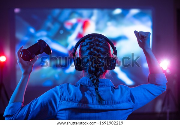 Girl in headphones plays a video game on the big\
TV screen. Gamer with a joystick. Online gaming with friends, win,\
prize. Fun entertainment. Teens play adventure games. Back view.\
Neon lighting