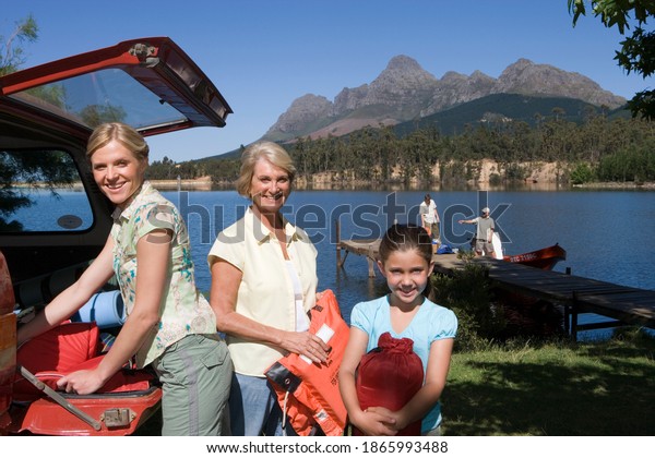 Girl helping her mother and grandmother\
unload life jackets from SUV with rest of the family on lake jetty\
beside a boat in the\
background.