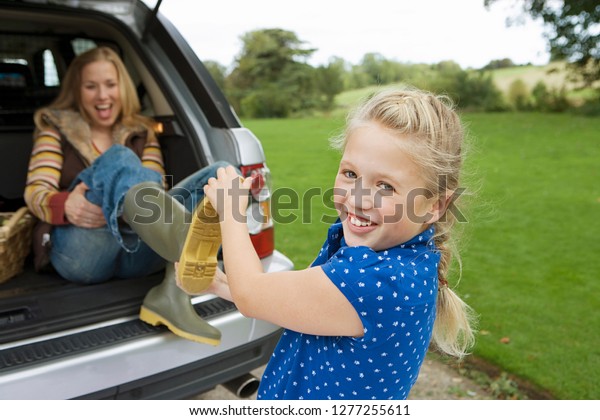 Girl helping mother\
with wellington boots in countryside as woman sits in trunk of car\
laughing and smiling