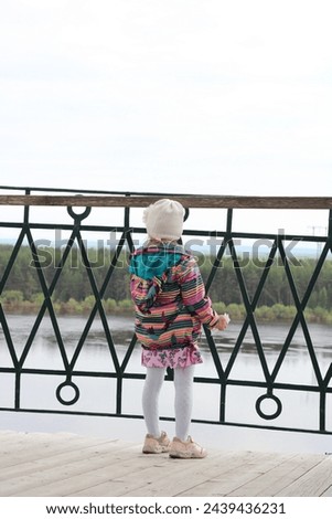 the girl with her back against the fence looks at the river in a bright jacket