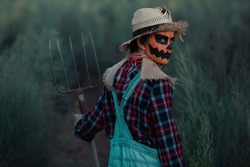 Girl With Her Back Turned Dressed As A Scarecrow With Halloween Make-up In A Green Orchard With A Pitchfork In Her Hand As She Holds It And Looks Back At It