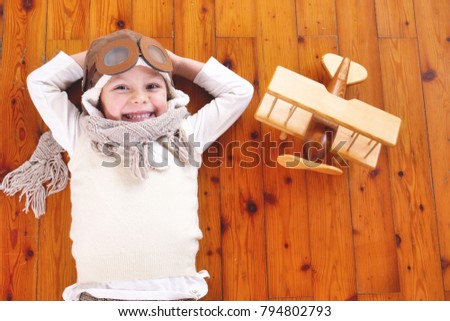A girl in her bedroom, dressed as a pilot or aviator, plays with her wooden airplane as she dreams with open eyes and thinks about the future. Concept of: success, future, dreams and work.