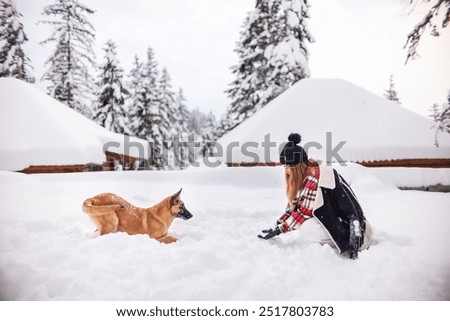 A girl and her dog enjoy a playful moment in deep snow, surrounded by snow-covered cabins and trees.