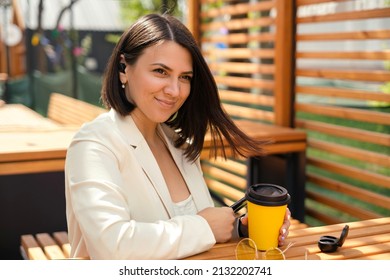 A Girl With Her Hair Fluttering In The Wind Sits Thoughtfully In A Street Cafe, Listening To Music Using In-ear Headphones. Coffee Break During Lunch. A Business Woman At A Table In A Publiс Place