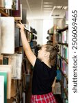 a girl with her hair in a ponytail chooses a book from a shelf in the library. She stands sideways to the camera, looking at the books around her with interest. The background is filled with tall shel