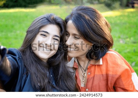 girl and her mother, beaming with smiles, take a cheerful selfie in a public park using a mobile phone, embracing the essence of love and happiness in a shared outdoor space