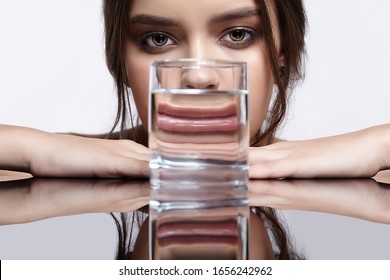 Girl Hides Her Face Behind A Glass With Water. Optical Distortion  Portrait Of Young Woman At The Mirror Table. Female On Gray Background.