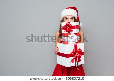 Girl hiding behind christmas gifts. Santa's helper. Dress and Santa's hat
