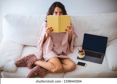 Girl Hiding Her Face Behind A Book While Sitting On The Sofa With A Laptop And Yogurt Or Ice Cream With Fresh Fruits In A Jar - Young Woman Relaxing And Reading In Cozy Room - Hardcover Book Mockup