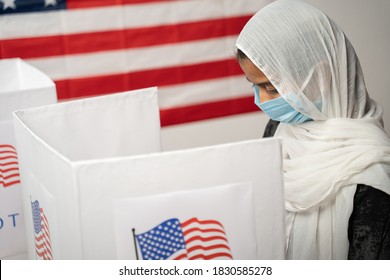 Girl In Hijab Or Head Covering And Mask Worn Busy At Polling Booth With US Flag As Background - Concept Of Voting In US Election