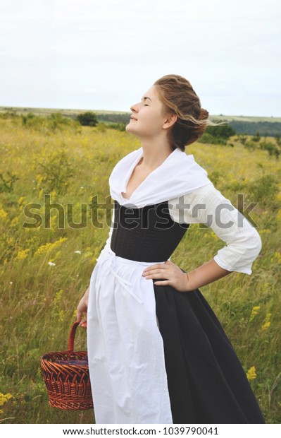 A\
girl in a historical costume with a basket standing in the field of\
yellow flowers. 18th century costume of a peasant\
girl