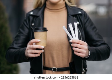 Girl holding coffee in a paper cup and sticks with sugar on her face. Hands of a girl holding hot coffee on the background of the mall.