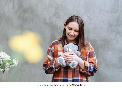 Girl holding a cute teddy bear toy. Woman in a checkered shirt on a background of gray concrete wall. Yellow spot and spring flowers on the wall. The brunette looked down.