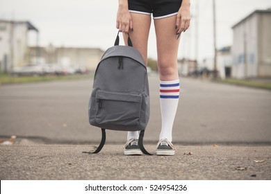 Girl Holding Generic Backpack Wearing Black Shorts And Tube Sock With Tennis Shoes Standing On Vacant Street. 