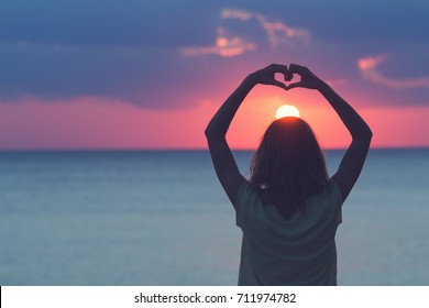 Girl holding a heart shape for the ocean / sea sunset.