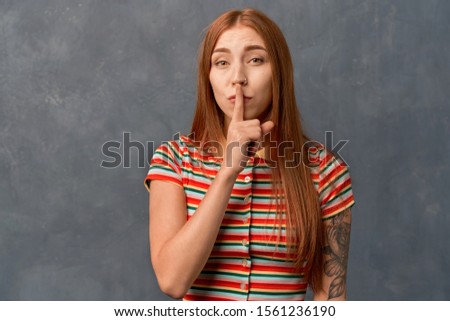 girl holding index finger at lips, asking to keep silence or not tell anyone secret, raising brows. Beautiful young girl with ginger long hair and tattoo on arm, wearing striped top, isolated on wall