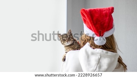 girl holding on shoulder tabby cat striped brown gray kitty female 5 month age cute adorable animal.woman back view blonde hair wearing christmas santa claus cap happy new year isolated wall window
