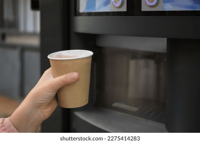 Niña sosteniendo una taza de papel con una bebida cerca del café y la máquina expendedora de bebidas calientes, armario. Espacio para texto