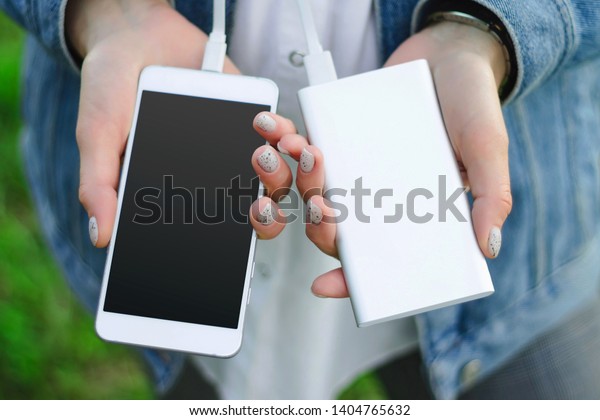 Girl\
holding power bank and a smart phone on green grass background.\
Girl charges her smartphone using power\
bank.