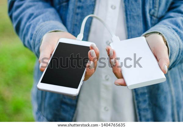 Girl\
holding power bank and a smart phone on green grass background.\
Girl charges her smartphone using power\
bank.