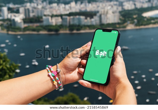 Girl holding smartphone with Bolt\
mobility company app on screen. City and bay with some boats in the\
background. Rio de Janeiro, RJ, Brazil. May\
2022.